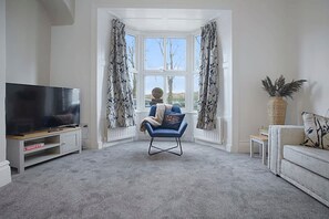 A living room with a flat-screen TV on a stand, a blue armchair, a small table, and a large potted plant. - Spacious Retreat With Backyard Bliss Sleeps 7 (Bishop Auckland)