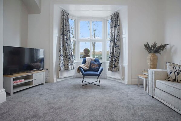 Sala de estar con televisor de pantalla plana sobre un mueble bajo, un sillón azul, una mesita y una planta grande en maceta.