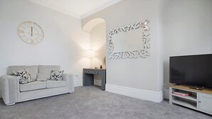 A living room with a grey sofa, a flat-screen TV, a decorative mirror, and a wall clock. - Spacious Retreat With Backyard Bliss Sleeps 7 (Bishop Auckland)