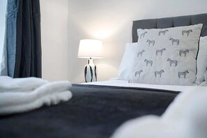 Una cama bien hecha con un cabecero oscuro y ropa de cama blanca, una lámpara de noche y un cojín con estampados de animales.