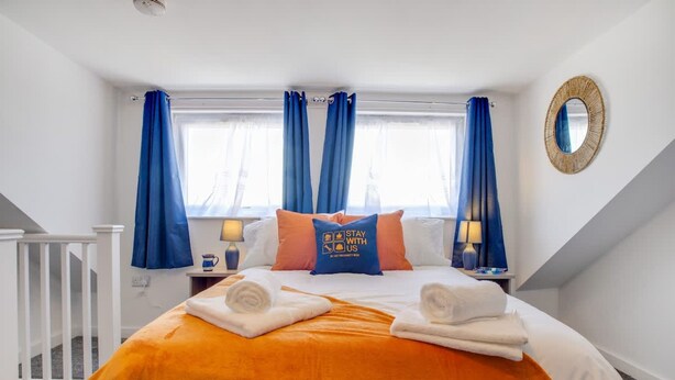A hotel room with a bed, two nightstands, a window with curtains, a mirror on the wall, and a white railing. - Central Darlington Sleeps 5 For Work or Leisure (Darlington)