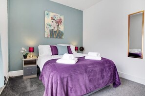 A hotel room with a bed, two bedside tables, a nightstand, a wall-mounted mirror, and a painting. - Central Darlington Sleeps 5 For Work or Leisure (Darlington)