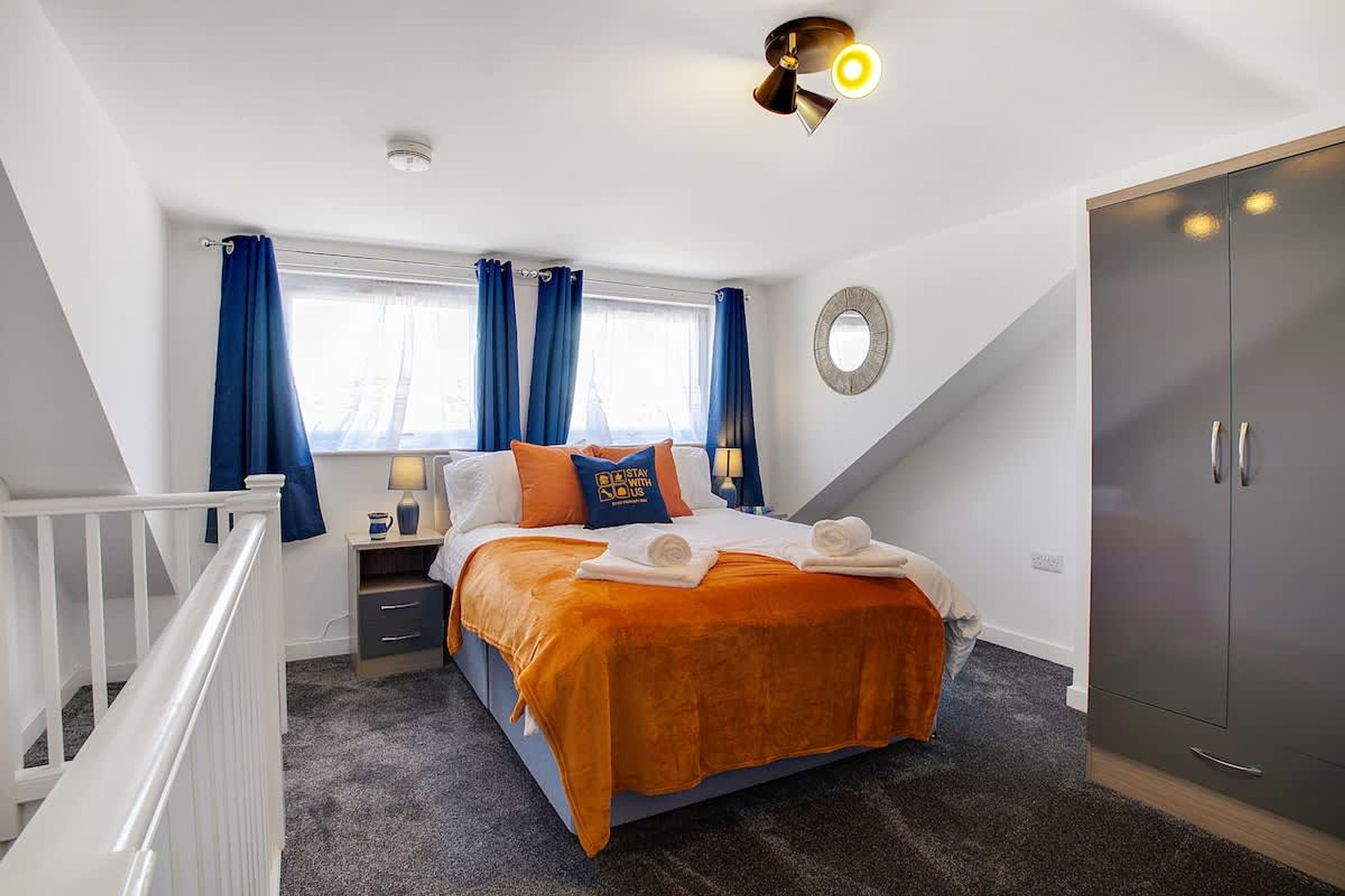 Um quarto com cama, mesa de cabeceira, luminária, guarda-roupa e janela com cortinas.