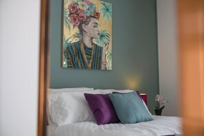 Ein Bett mit weißen Laken und zwei Kissen, eines violett und eines türkis, in einem Zimmer mit türkis gestrichener Wand und einem Gemälde über dem Bett.