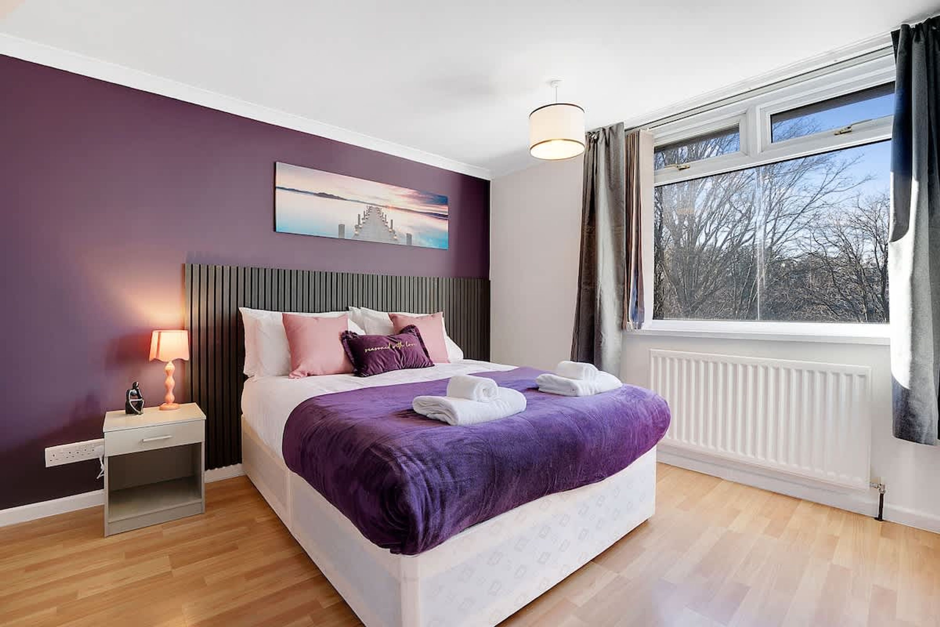 A bedroom with a bed, a nightstand, a lamp, a window with curtains, and a painting on the wall.