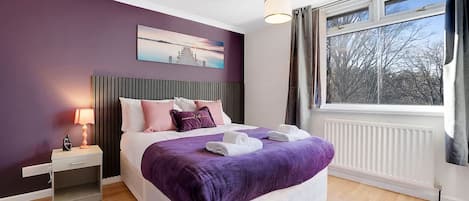 A bedroom with a bed, a nightstand, a lamp, a window with curtains, and a painting on the wall.