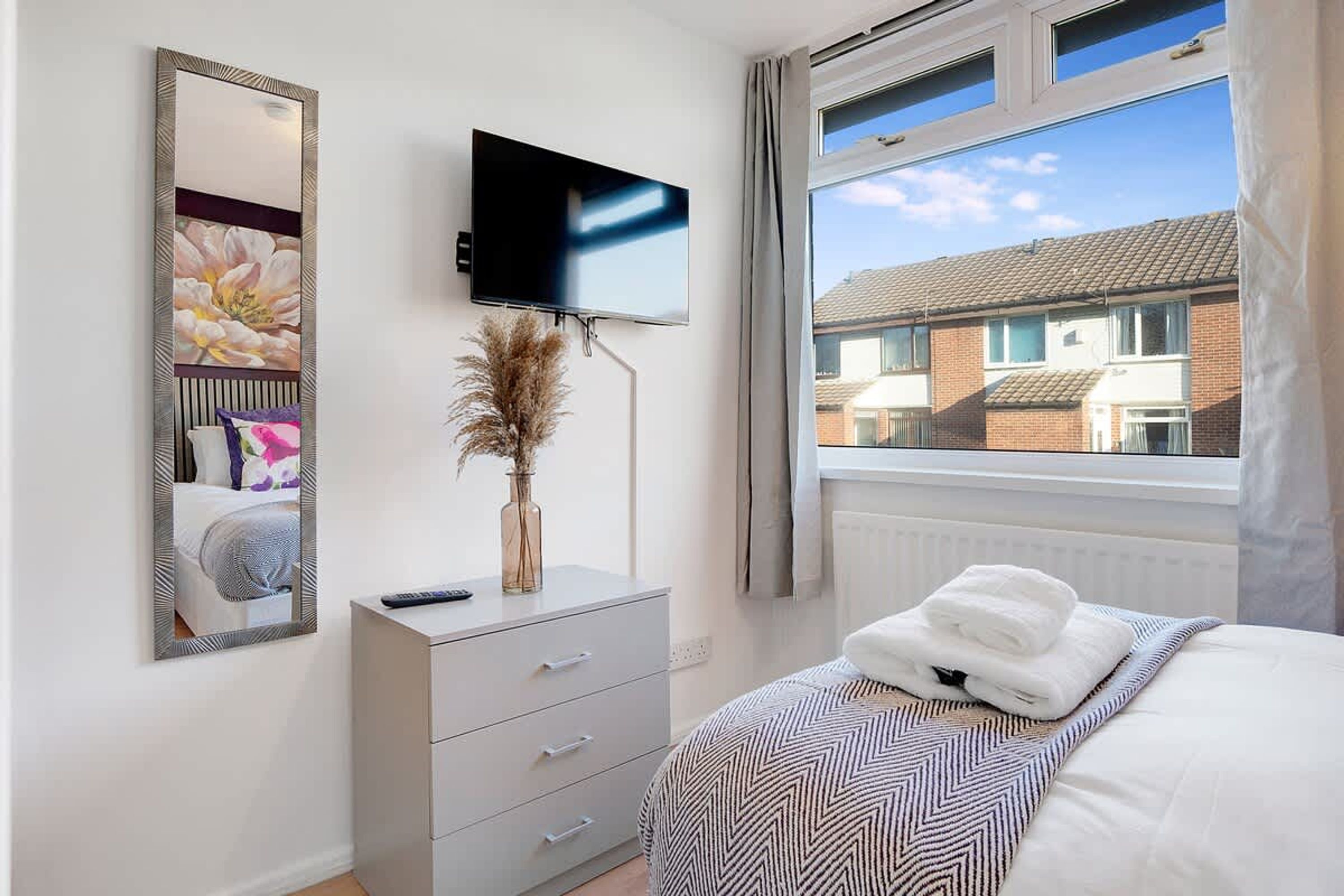 Un dormitorio con cama, cómoda, televisor montado en la pared y ventana con vista a casas vecinas.