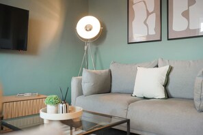 Ein modernes Wohnzimmer mit einem grauen Sofa, einem Glastisch, einer Stehlampe und abstrakter Wandkunst.