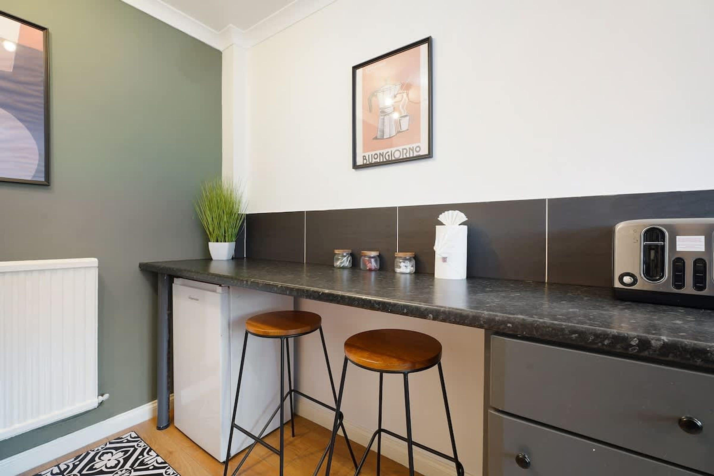 A kitchen with a countertop, two stools, a refrigerator, and a coffee machine.