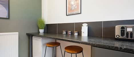 A kitchen with a countertop, two stools, a refrigerator, and a coffee machine.