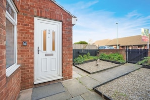 A residential property with a white door, brick walls, a small garden with gravel and plants, and a black fence. - Charming 3 Bedroom Home Near Coast Town (Redcar)