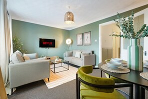 Ein Wohnzimmer mit einer Couch, einem Couchtisch, einem Fernseher mit der Anzeige 'NETFLIX' und einem Essbereich mit Tisch und Stühlen.