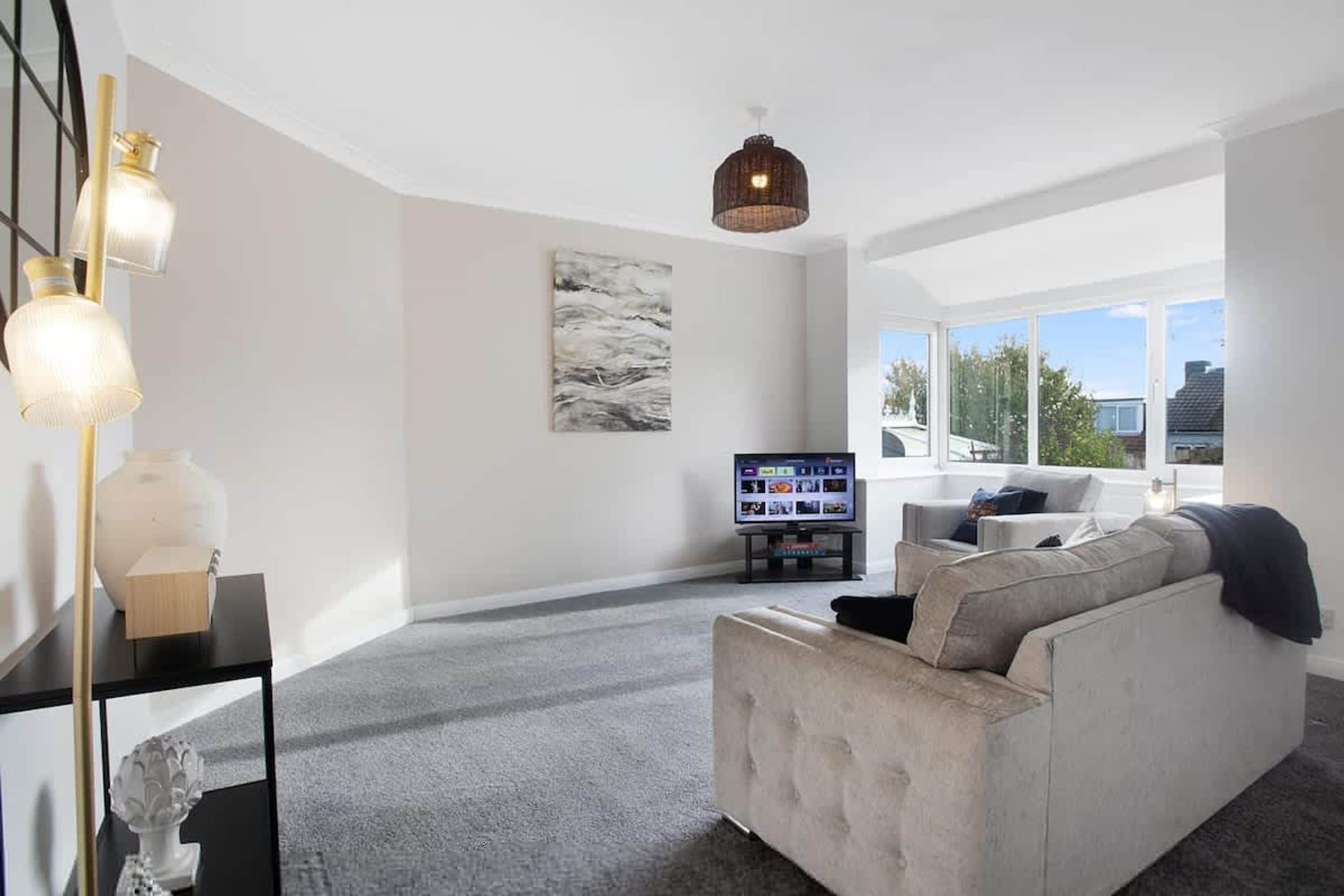 Ein modernes Wohnzimmer mit einem grauen Sofa, einem Flachbildfernseher und einem großen Fenster mit Blick ins Freie.