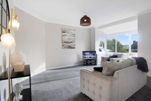 Ein modernes Wohnzimmer mit einem grauen Sofa, einem Flachbildfernseher und einem großen Fenster mit Blick ins Freie.