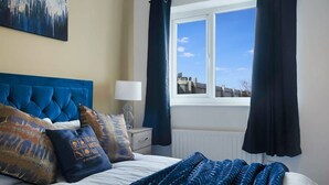 Une chambre avec un grand lit, une fenêtre avec des rideaux, une table de chevet avec une lampe et un tableau accroché au mur.