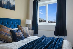 A bedroom with a large bed, a window with curtains, a nightstand with a lamp, and a painting on the wall. - Spacious Living Room and Modern Comforts Sleeps 4 (Peterlee)