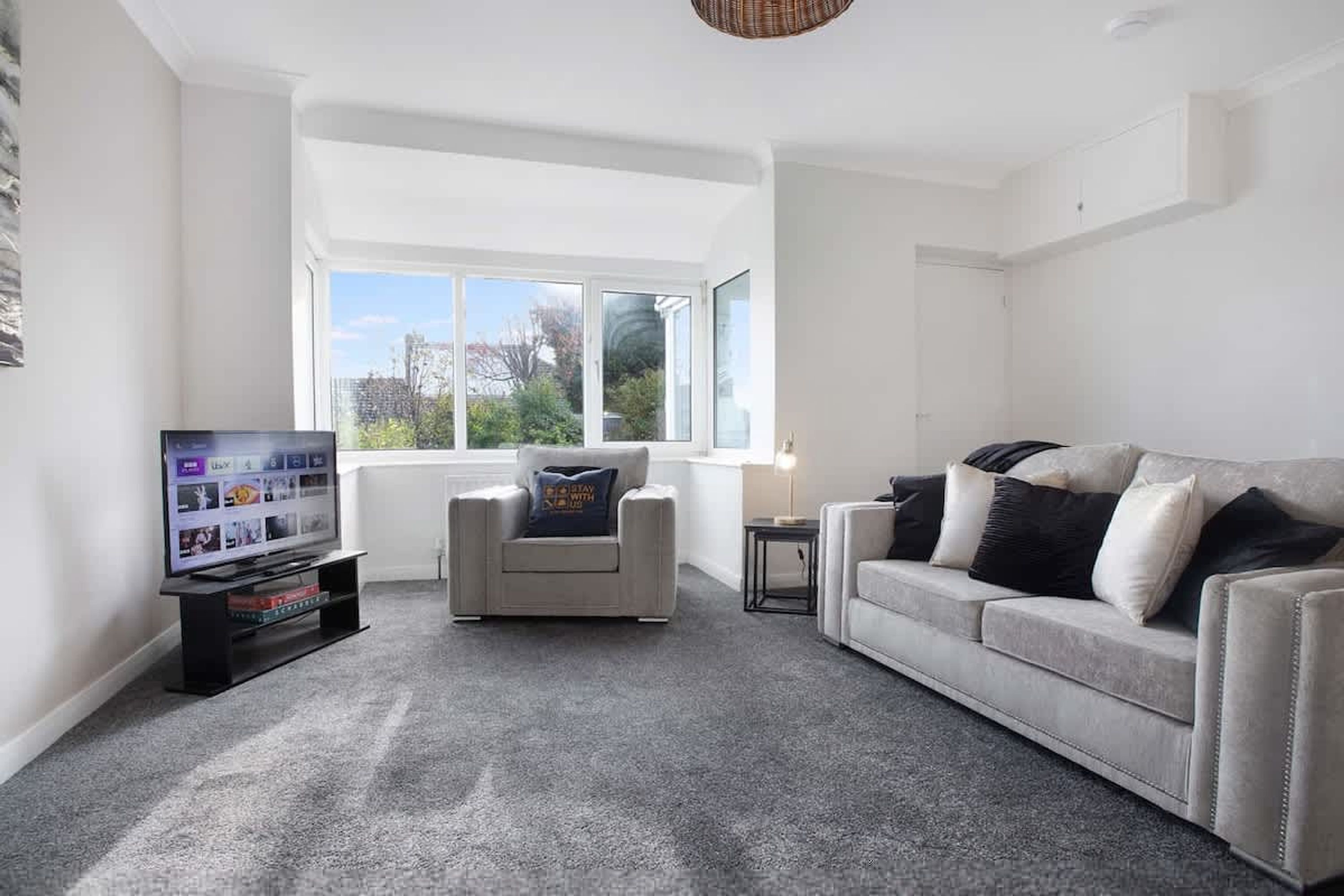 Una sala de estar moderna con alfombra gris, un sofá blanco, un sillón gris, un televisor sobre un mueble bajo y un ventanal grande.