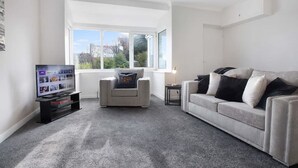 Un salon moderne avec un tapis gris, un canapé blanc, un fauteuil gris, un téléviseur posé sur un meuble et une grande fenêtre.