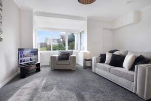 A modern living room with a grey carpet, a white sofa, a grey armchair, a TV on a stand, and a large window. - Spacious Living Room and Modern Comforts Sleeps 4 (Peterlee)