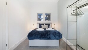 A bedroom with a bed, two wall-mounted pictures, a bedside table with a lamp, and a metal drying rack. - Sunderland 3BR Retreat Sleeps 6 by Stay With Us (Sunderland)