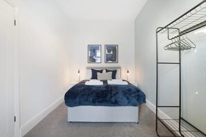 Ein Schlafzimmer mit einem Bett, zwei an der Wand befestigten Bildern, einem Nachttisch mit Lampe und einem Wäscheständer aus Metall.