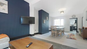 A modern living room with a flat-screen TV, a dining area with a table and chairs, and a kitchen visible through an open door. - 5BR Sleeps 10 Perfect for Contractors in Durham (Durham)