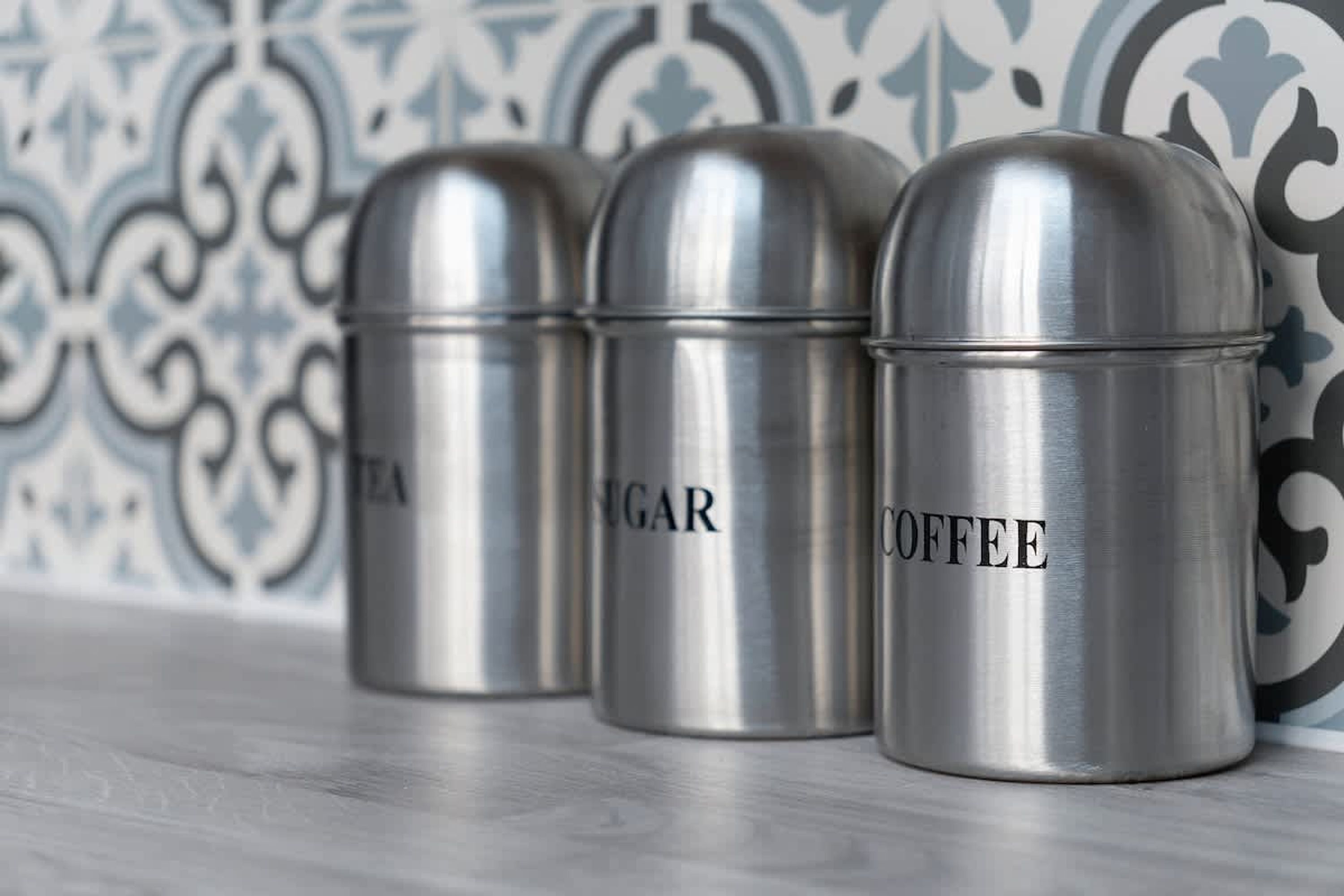 Three stainless steel containers labeled tea, sugar, and coffee on a wooden surface with a patterned wall in the background.