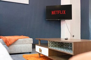 A modern living room with a flat-screen TV mounted on the wall displaying 'NETFLIX', a grey sofa with an orange throw blanket, and a wooden side table with a patterned glass top. - 5BR Sleeps 10 Perfect for Contractors in Durham (Durham)