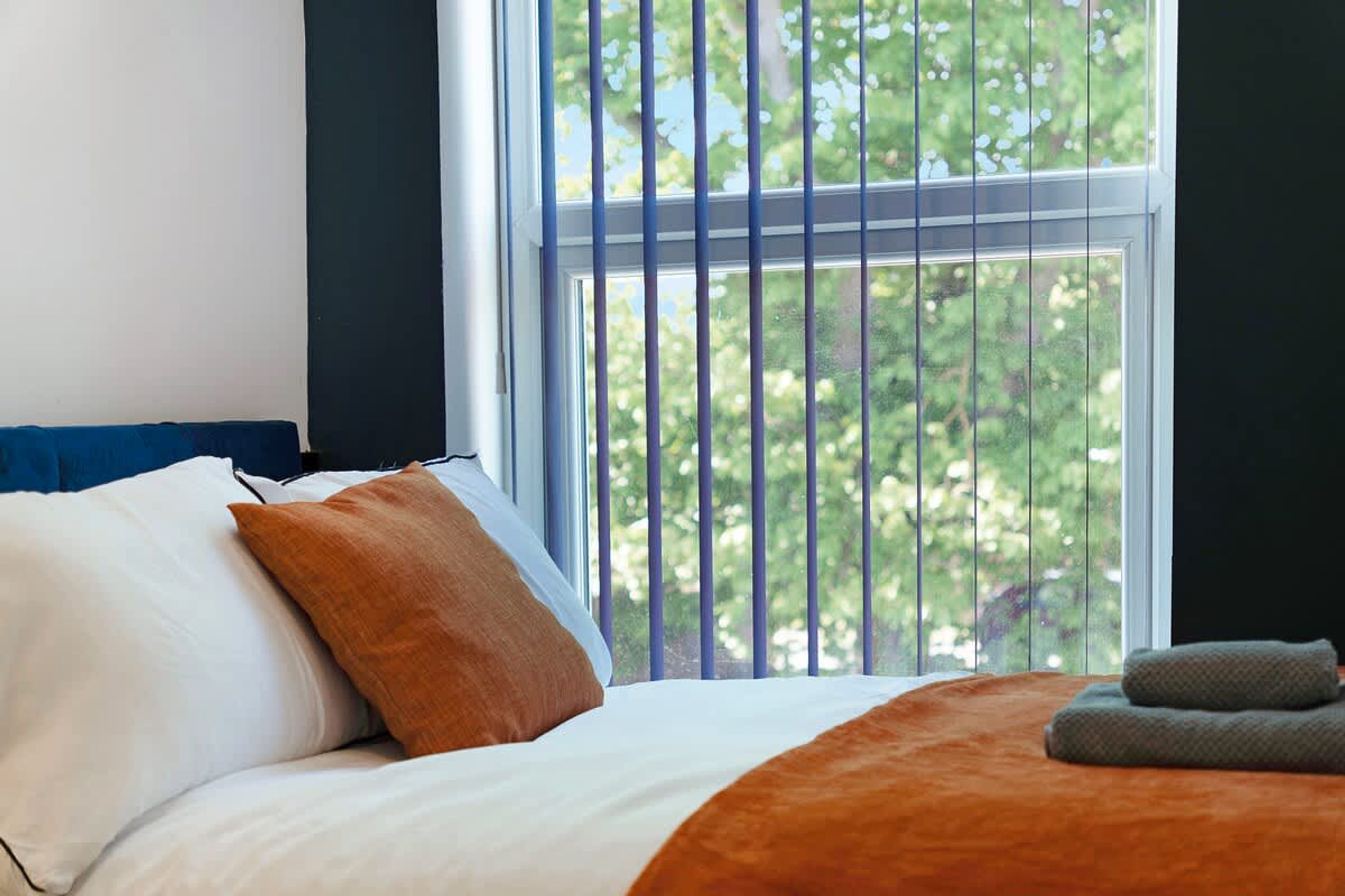 A bed with white and orange bedding, a window with vertical blinds, and a view of trees outside.