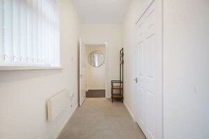 A narrow hallway with white walls, a radiator, a window with blinds, a door with a mirror, and a shelf with items.