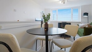 Et moderne spiseområde med et rundt bord, stole, en vase med en plante og udsigt til himlen gennem et vindue.