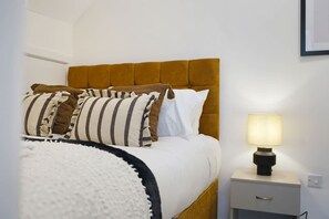 Ein ordentlich bezogenes Bett mit einem gestreiften Kopfkissen, einem weißen Kopfkissen und einem braunen Kopfteil. Ein Nachttisch mit einer Lampe und einer Schublade ist ebenfalls vorhanden.
