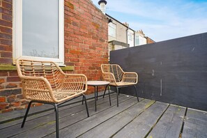 Eine Terrasse mit Korbstühlen und einem kleinen Tisch, eine Ziegelwand und ein schwarzer Zaun.