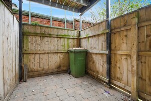 Un espacio exterior pequeño y cercado, con una valla de madera, un bote de basura verde y suelo de ladrillo.