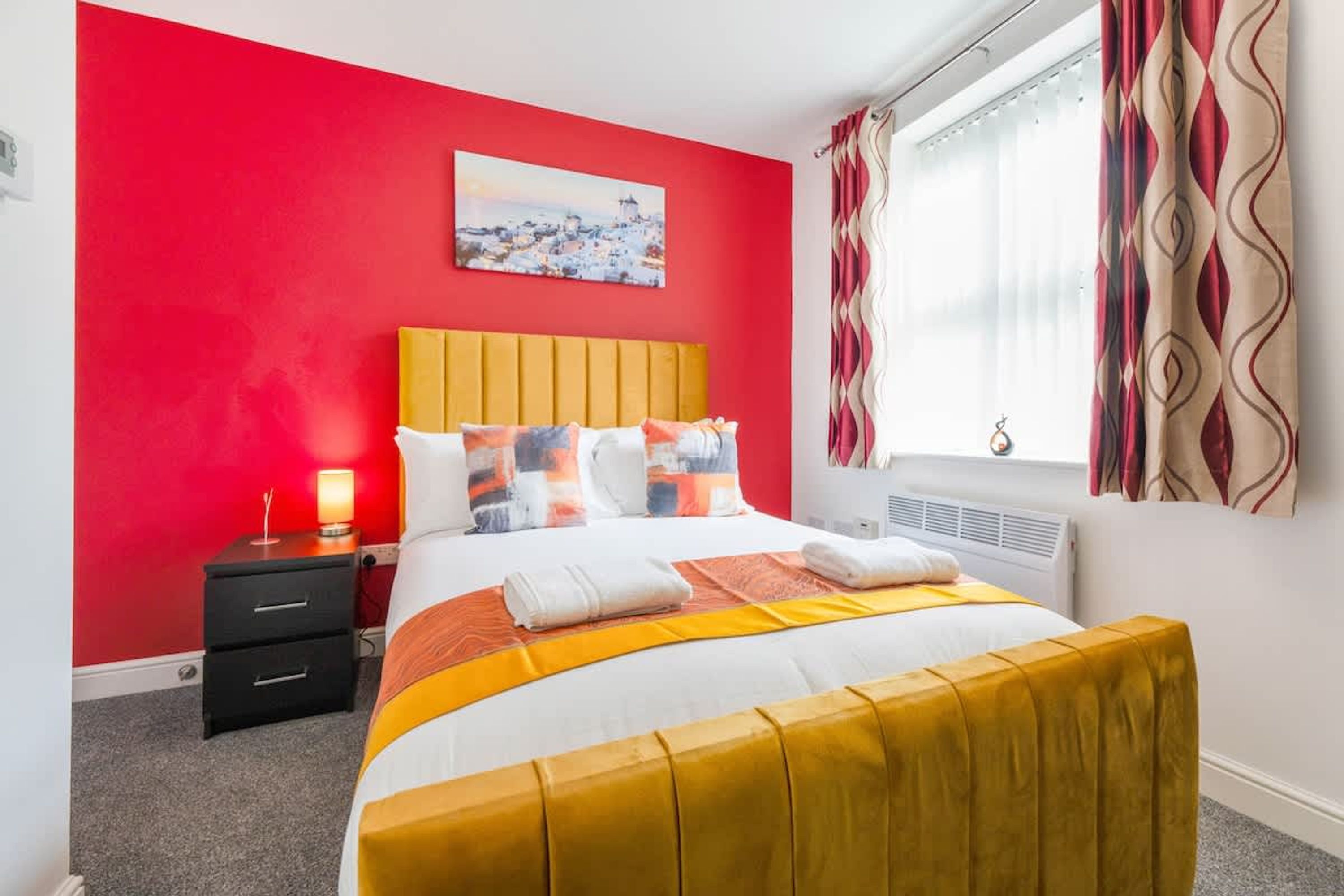 A hotel room with a red wall, a bed with a yellow headboard, and a window with patterned curtains.