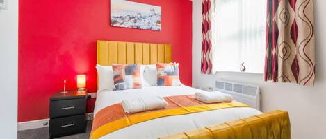 Una habitación de hotel con una pared roja, una cama con cabecera amarilla y una ventana con cortinas estampadas.