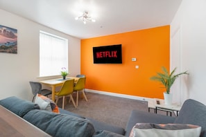 A modern living room with a large orange wall, a flat-screen TV mounted on it, a dining area with a table and chairs, and a sofa with cushions. - Stylish 5 Bed Flat in Thornaby Ideal for Groups (Stockton-on-Tees)