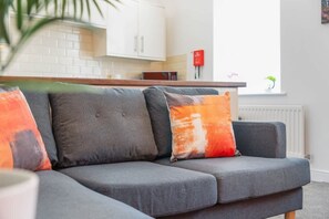 En grå soffa med två kuddar, ett vitt kök i bakgrunden och ett fönster med en växt.