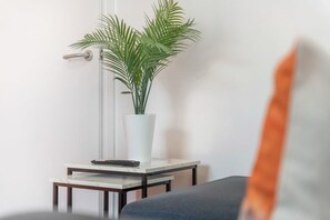 Ein Zimmer mit weißer Wand, einer Topfpflanze auf einem kleinen Tisch, einer Fernbedienung und einem Teil eines Sofas.
