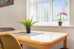 Ein Zimmer mit Holztisch, einem Stuhl, einem Blumentopf mit Farn und einem Fenster mit Blick auf Bäume und eine rosa Blume.
