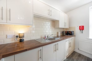 A modern kitchen with white cabinets, a wooden countertop, a built-in microwave, and a red fire blanket on the wall. - Stylish 5 Bed Flat in Thornaby Ideal for Groups (Stockton-on-Tees)