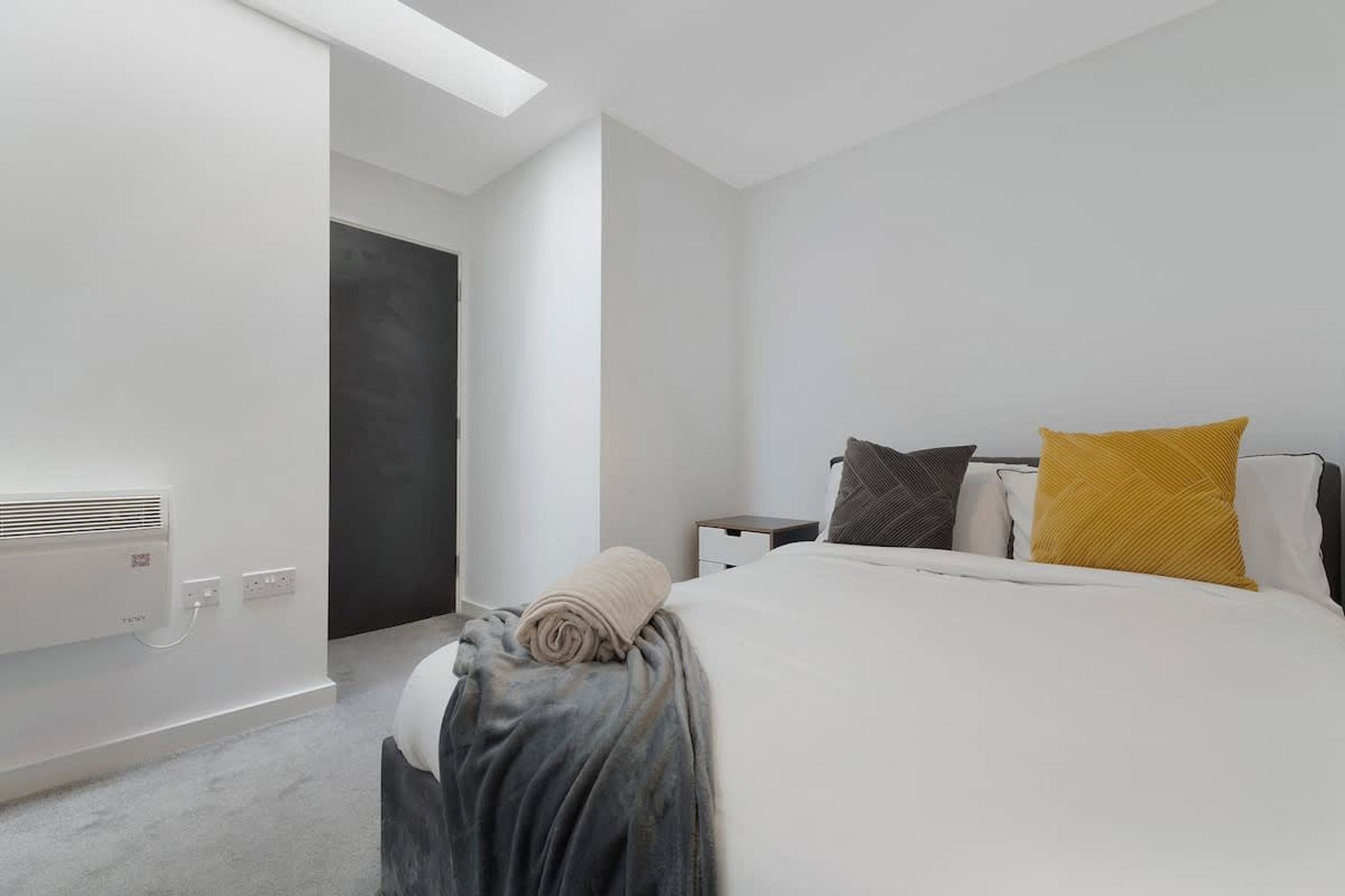 A modern hotel room with a bed, bedside tables, a nightstand, a rolled-up blanket, and a wall-mounted air conditioning unit.
