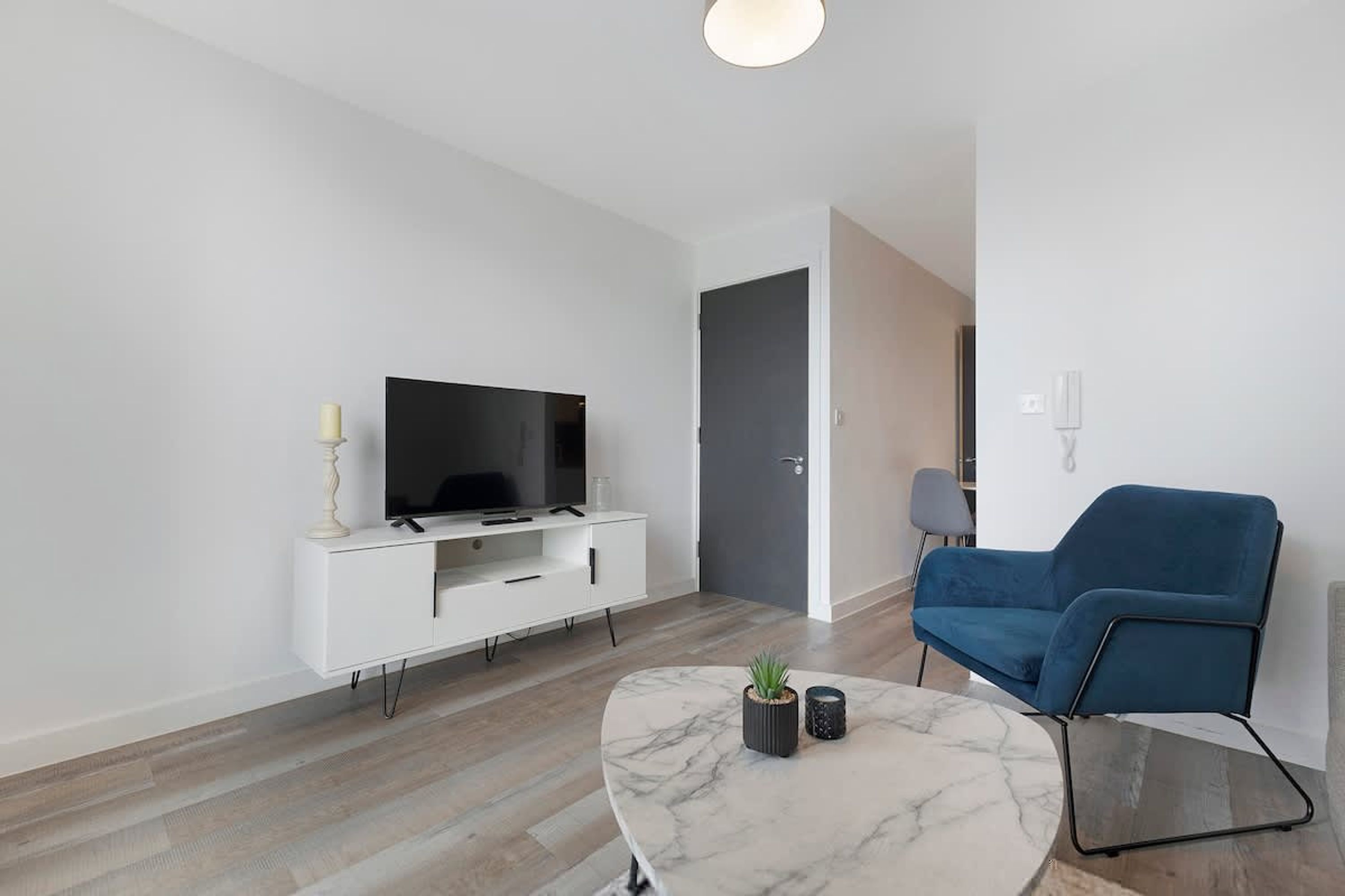 A modern living room with a flat-screen TV on a white TV stand, a blue sofa, a marble coffee table, and a small potted plant.