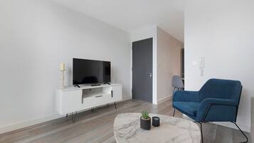 Een moderne woonkamer met een flatscreen televisie op een witte tv-meubel, een blauwe bank, een marmeren salontafel en een kleine potplant.
