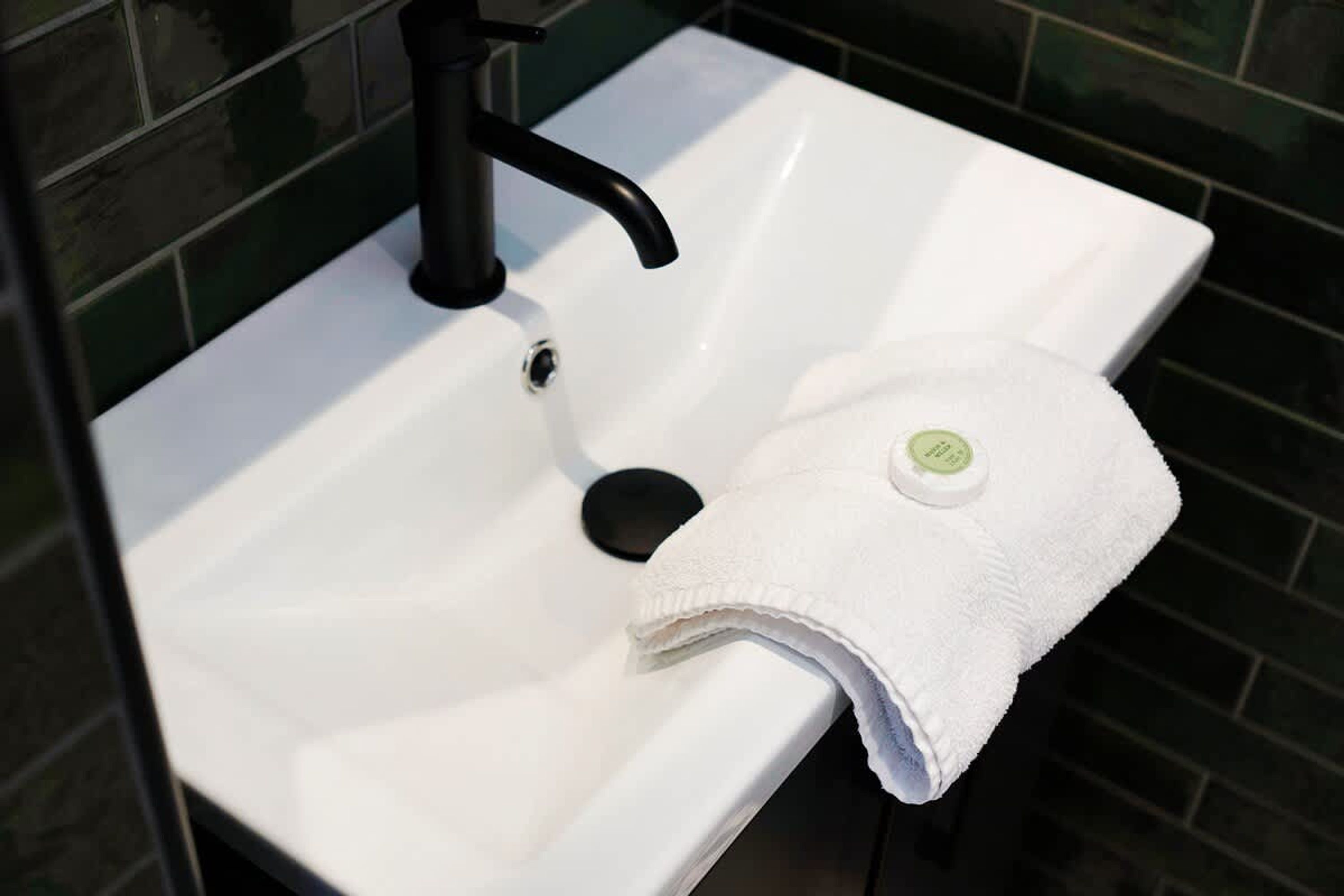 Ein weißes Waschbecken mit einem schwarzen Wasserhahn und einem weißen Handtuch mit grünem Logo an einer gefliesten Wand.