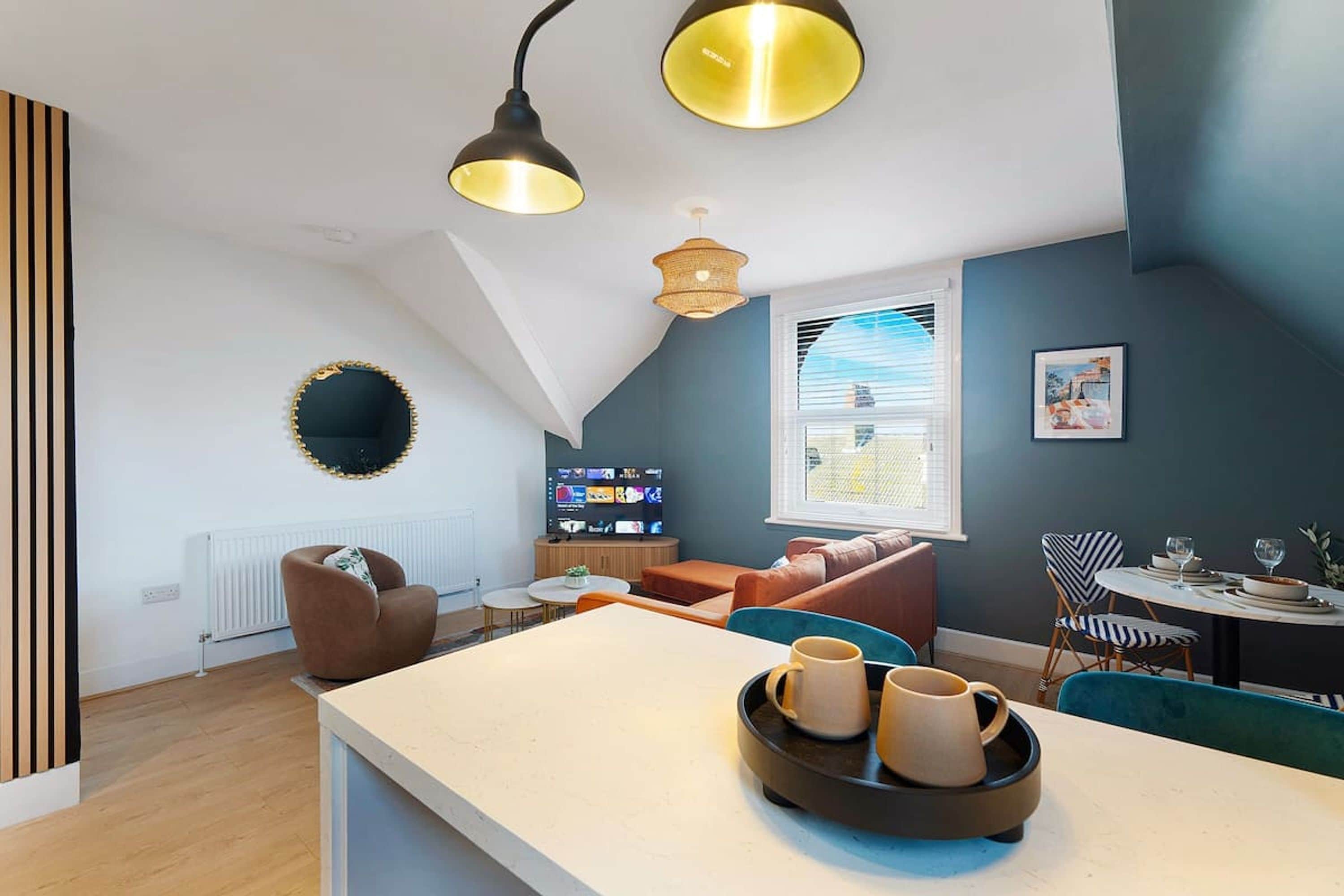 A modern living space with a loft-style ceiling, a television, a dining area, and a kitchen counter with mugs.