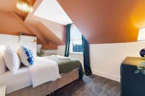 Ein Schlafzimmer mit Bett, Kissen, Nachttisch, Lampe und einem Fenster mit Vorhängen.