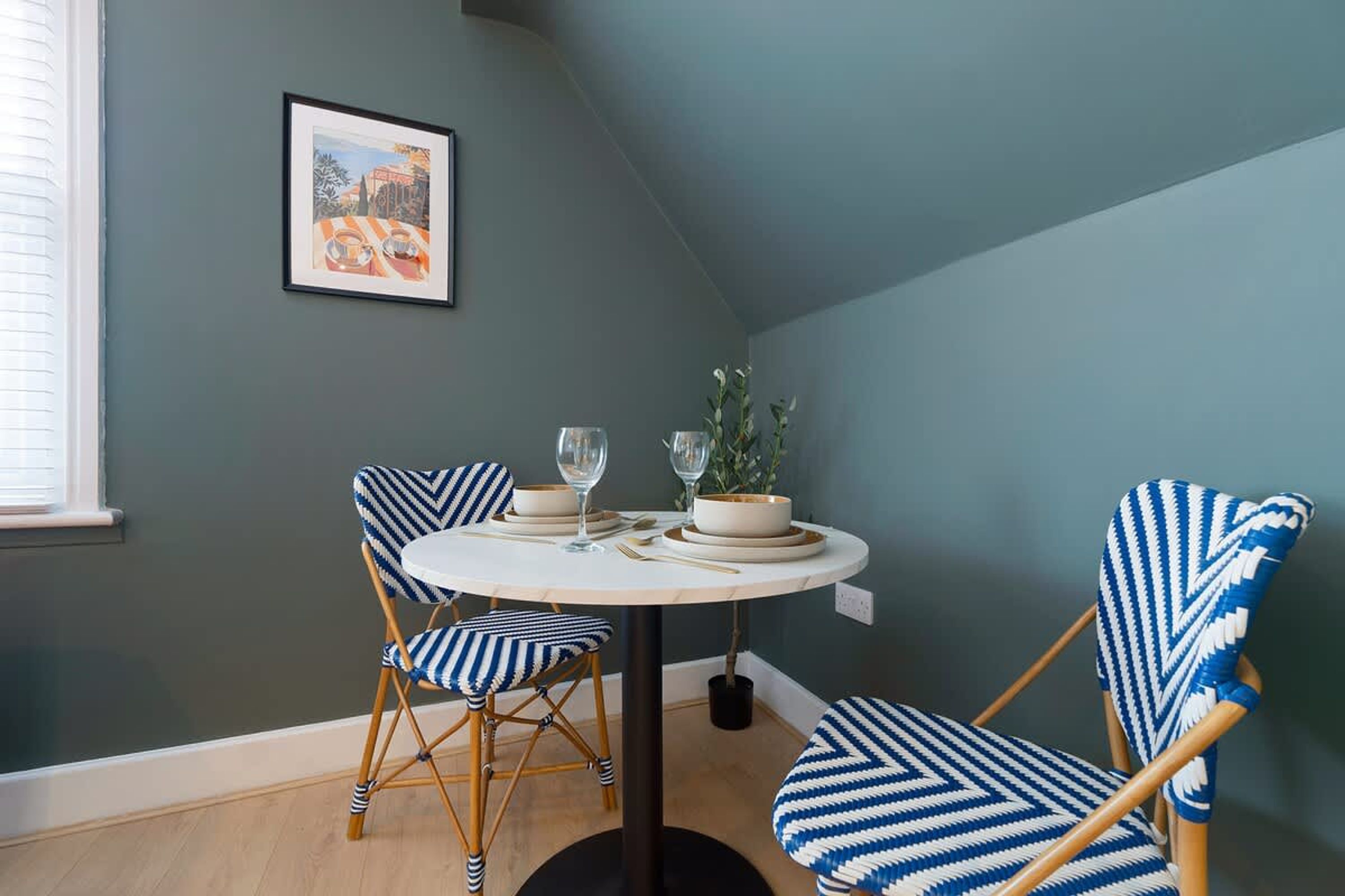 A dining area with a round table, two chairs, a potted plant, and a framed picture on the wall.