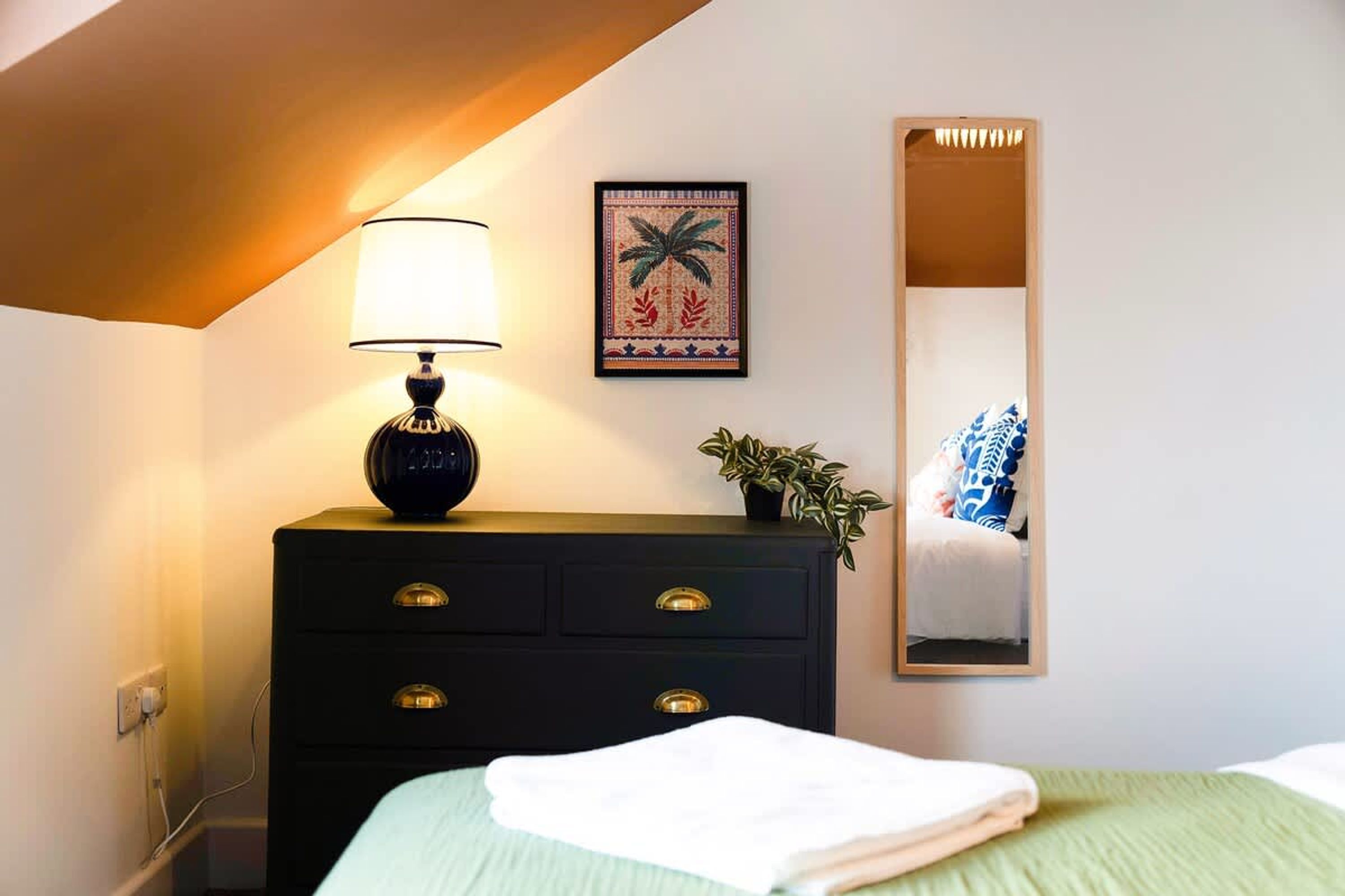 A bedroom with a bed, a dresser with drawers, a lamp, a framed picture, a potted plant, and a mirror.