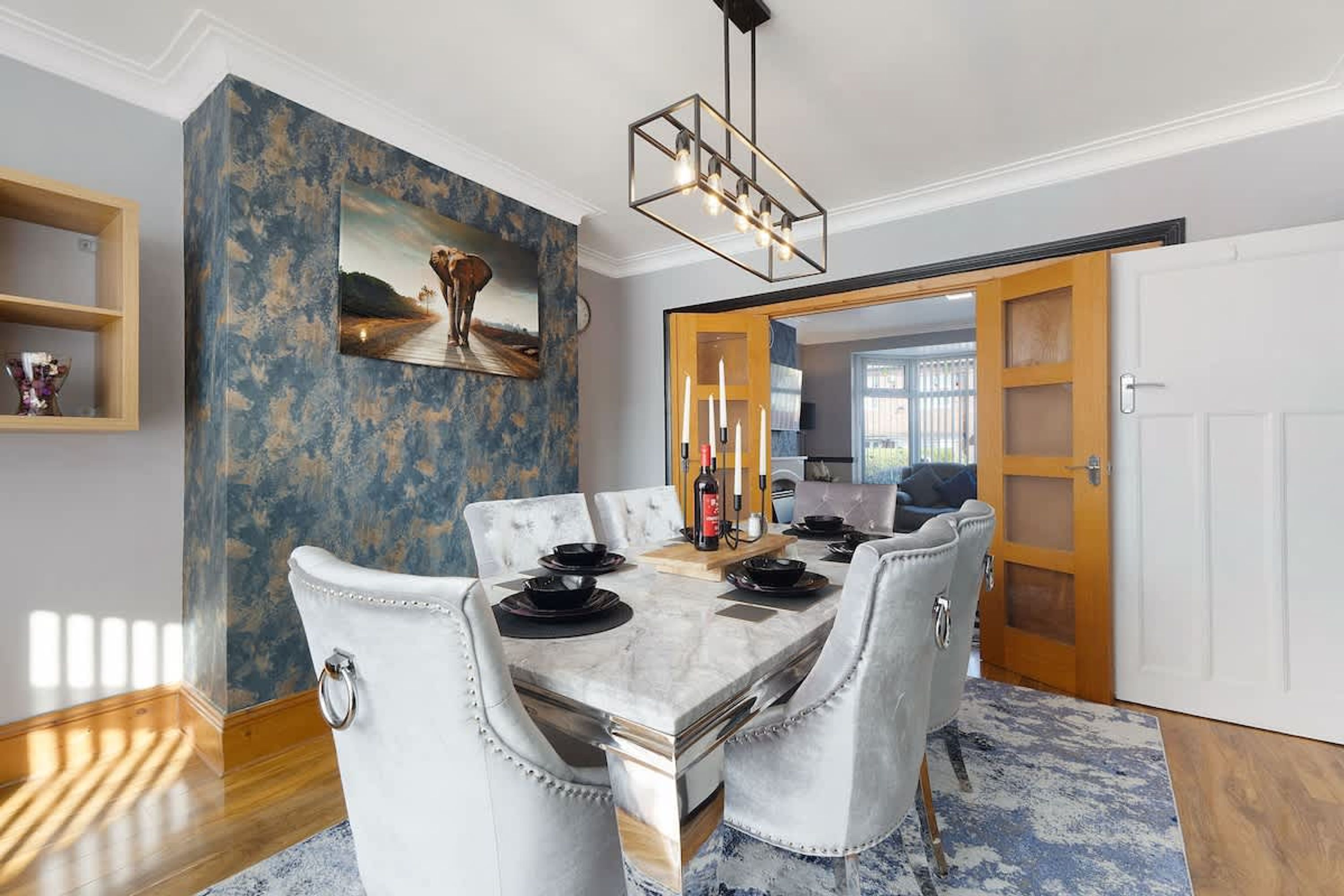A dining area with a marble table, white upholstered chairs, a chandelier, and a large abstract painting on the wall.